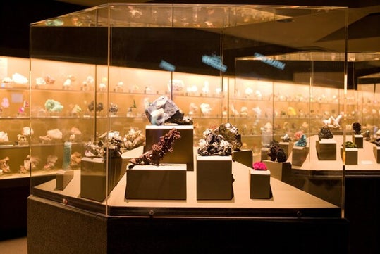 A display case in a museum features various mineral specimens on pedestals, with more minerals visible in the background. The area is well-lit, highlighting their colors and textures.