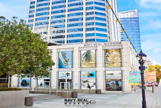 Exterior view of the Navy SEAL Museum in San Diego, featuring large photos in windows, nearby trees, a lamppost, and tall buildings in the background.