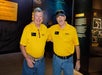 Two men in yellow polo shirts and jeans stand side by side, smiling at the camera, in a museum exhibit with informational displays in the background.