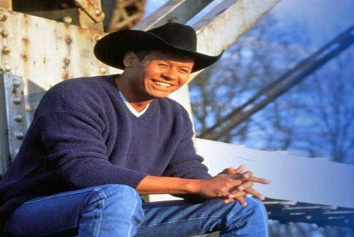 A man in a black cowboy hat and blue sweater smiles while sitting outdoors on a metal structure, with trees and blue sky in the background.