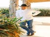 Man in a white shirt, jeans, cowboy hat, and boots leans against a wooden post outdoors near green plants on a sunny day.