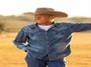 A man wearing a cowboy hat, denim shirt, and jeans stands outdoors in a sunlit field, smiling and looking to the side.