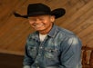 A man wearing a black cowboy hat and denim shirt sits on a wooden chair, smiling, with a wooden panel background.