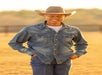 A man wearing a denim shirt, jeans, and a cowboy hat stands outdoors in a sunlit field, smiling with his hands in his pockets.