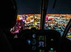 Night view of a city skyline with brightly lit buildings seen through the cockpit of a helicopter, showing illuminated flight instruments.