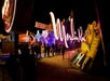 People stand in line at night in front of large, illuminated Moulin Rouge and other vintage neon signs under colorful lights.