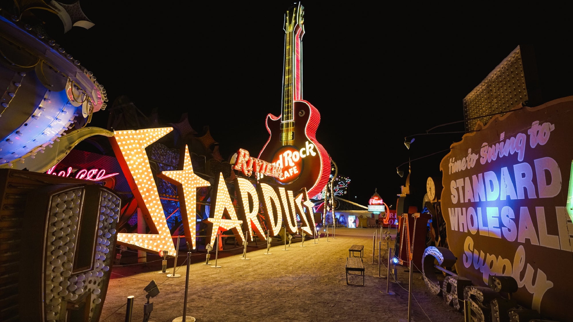A nighttime view of vintage neon signs, including a large "Stardust" sign and a giant guitar for the Hard Rock Cafe, displayed outdoors at a neon museum.