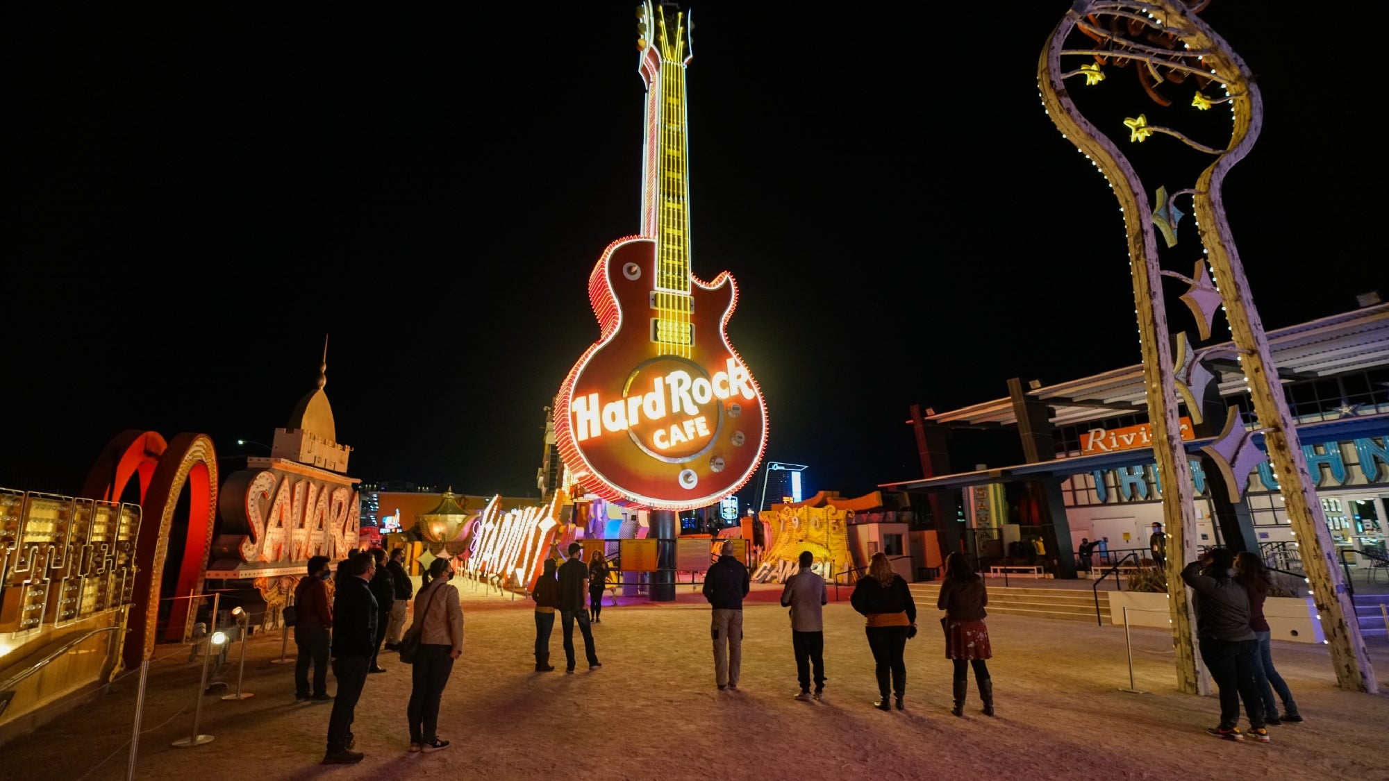 People stand in front of a large illuminated Hard Rock Cafe guitar sign at night in an outdoor neon sign display area.