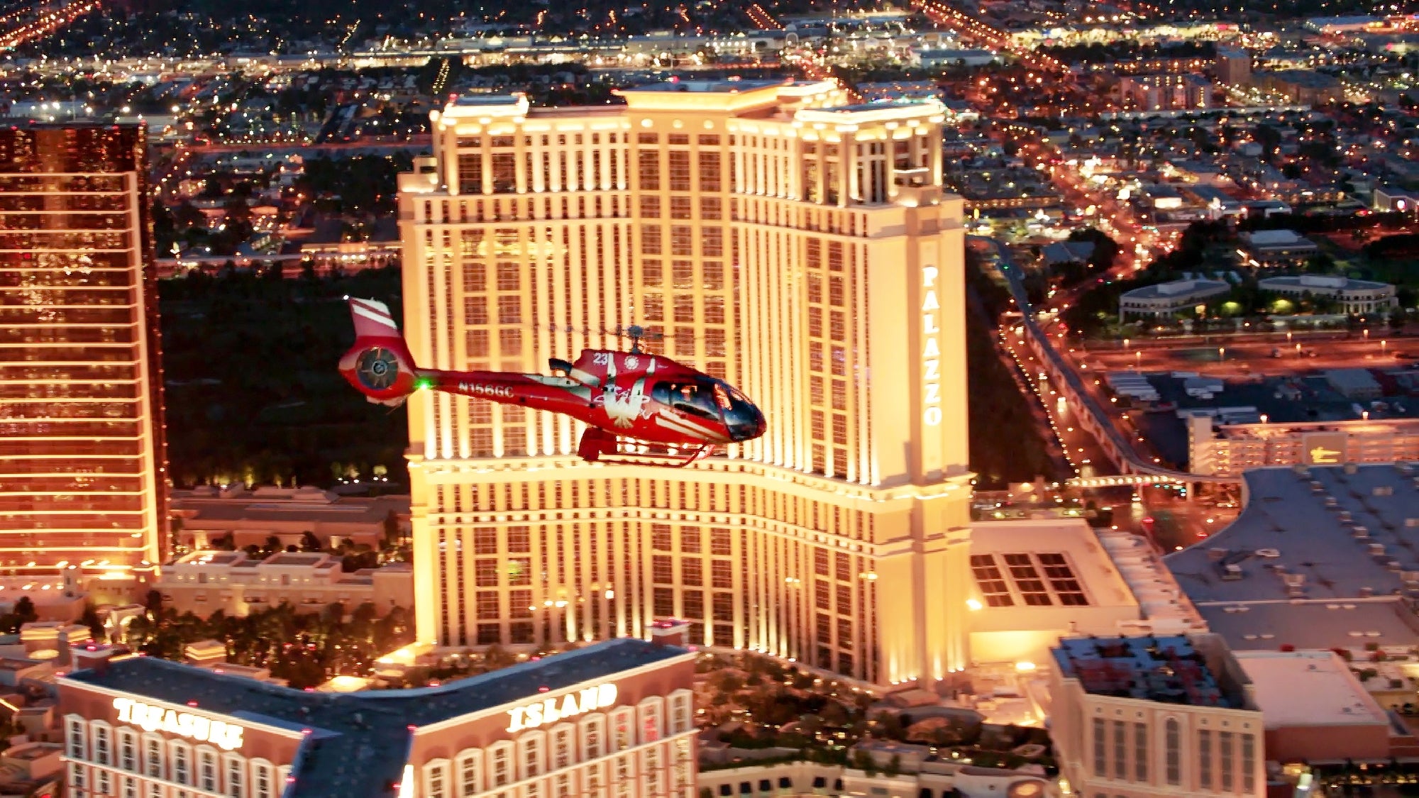 A red helicopter flies in front of a tall, illuminated hotel building labeled "Palazzo" in a brightly lit cityscape at dusk or night.
