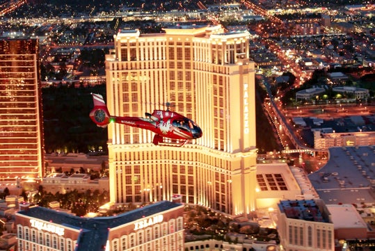 A red helicopter flies in front of a tall, illuminated hotel building labeled "Palazzo" in a brightly lit cityscape at dusk or night.