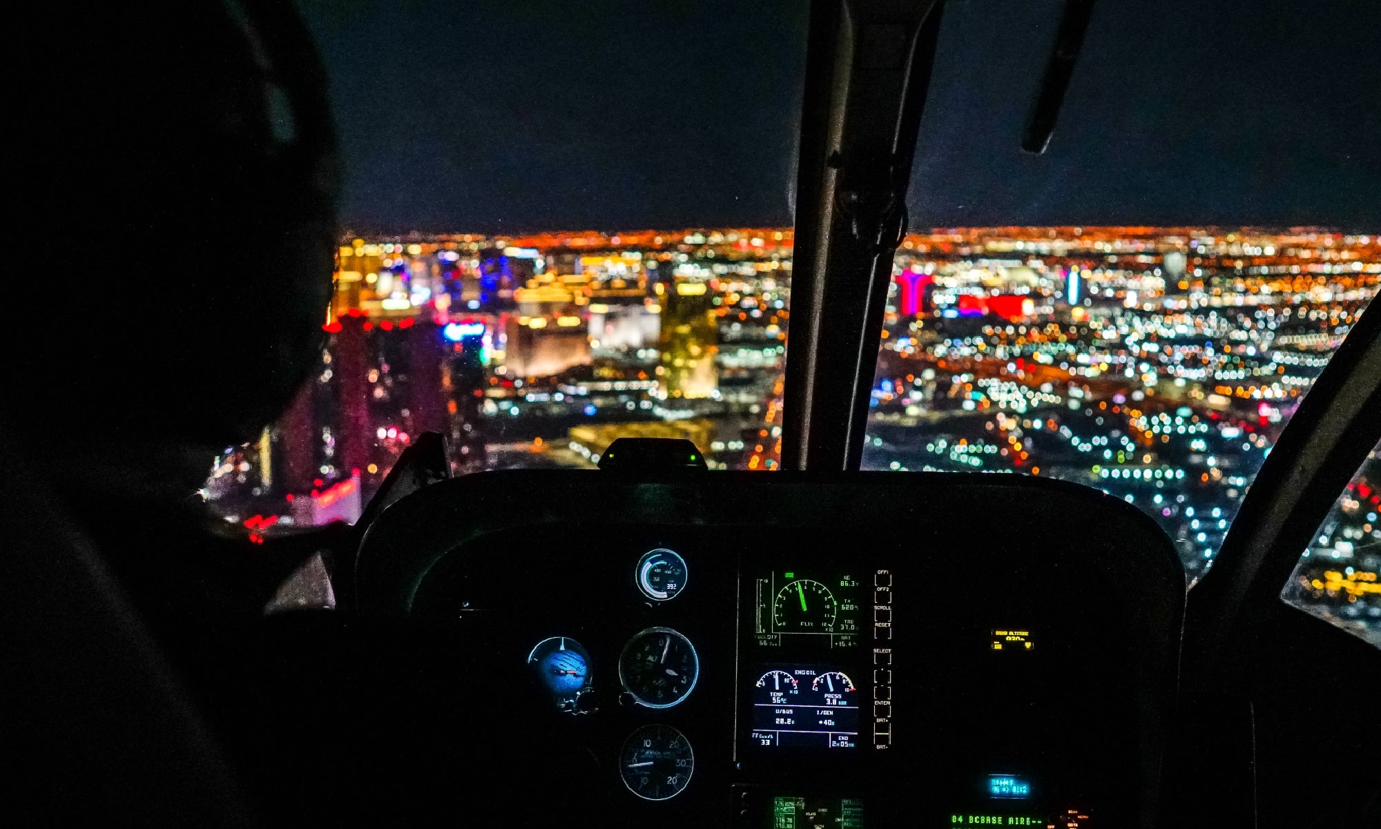 View from inside a helicopter cockpit at night, with illuminated instrument panels in the foreground and a brightly lit cityscape in the background.