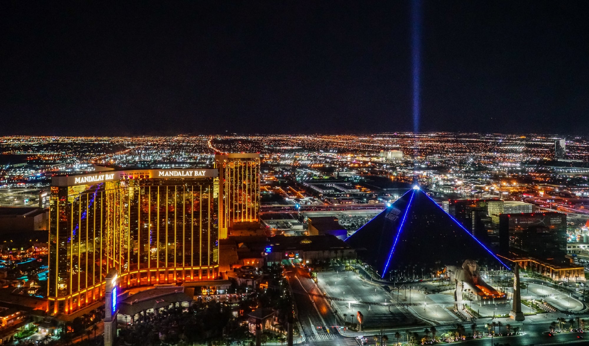 A nighttime aerial view of the Las Vegas Strip, featuring the illuminated Mandalay Bay hotel and the Luxor pyramid with a bright beam of light shining into the sky.