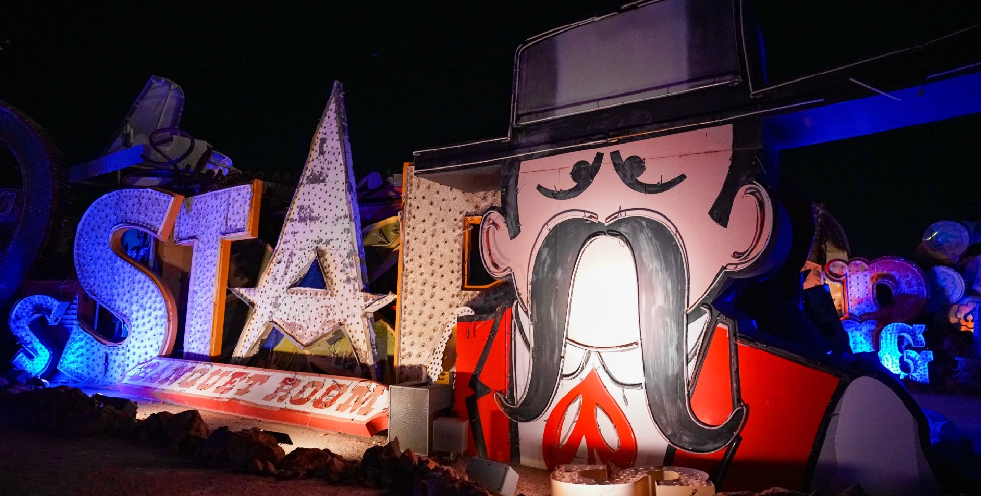 Large, illuminated cowboy face with a mustache and hat stands next to retro "STAR" sign at night among other vintage neon signage.