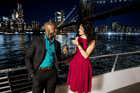 A man and woman talking and cheering their wine glasses together near the railing of a cruise boat with NYC in the background at night.