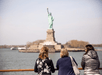 Three women leaning on the railing of a cruise boat while looking at the Statue of Liberty in the distance on a sunny day.