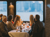 A group of people seated at a white cloth covered table with food and drinks in front of them, all turned to look out the window at the end of the table.
