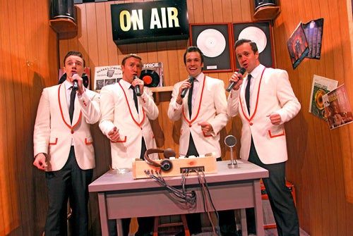 Four men in matching white blazers with orange trim sing into microphones in a retro radio studio with an "ON AIR" sign and records on the wood-paneled walls.