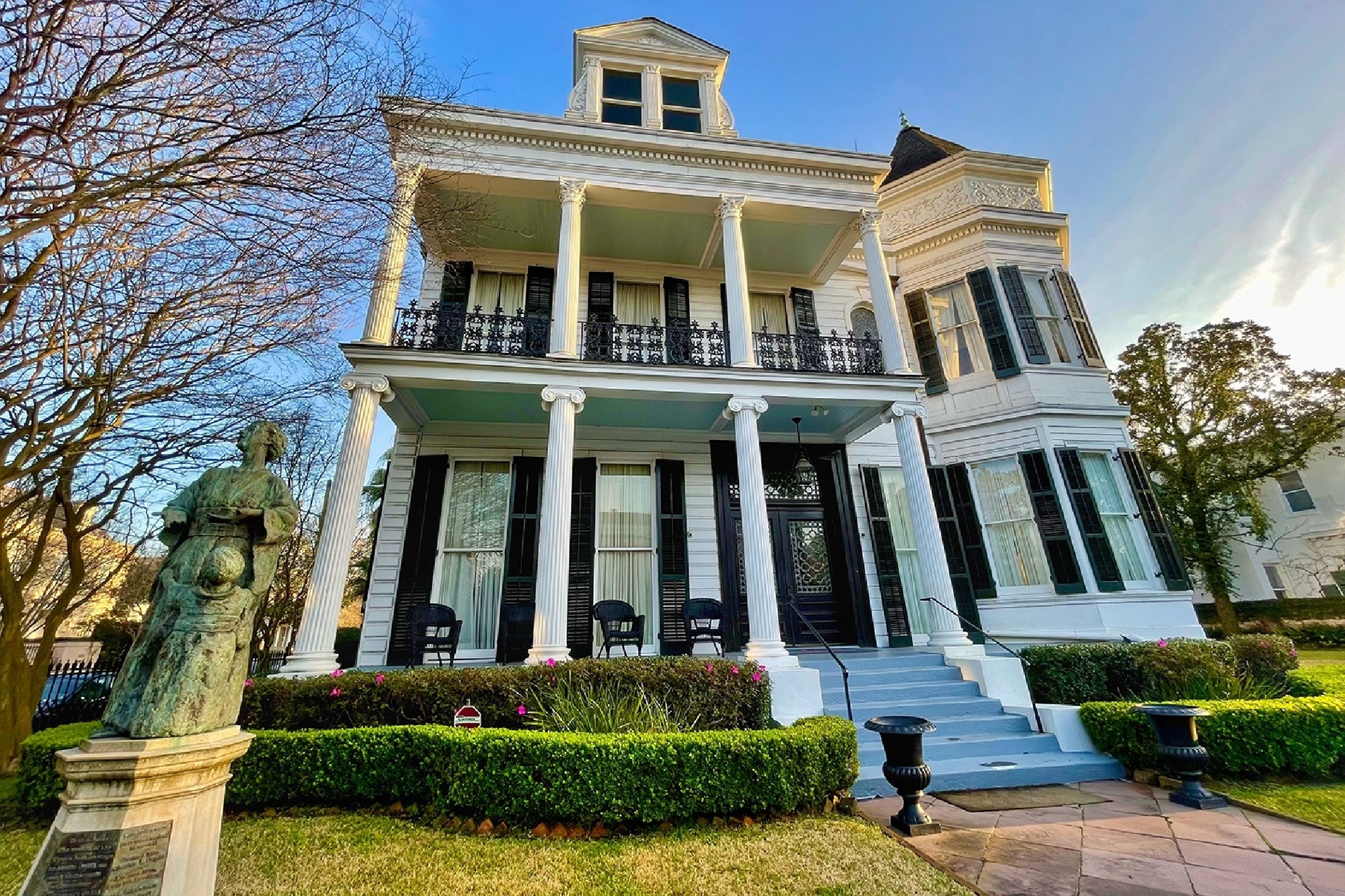 A large white two-story historic house with tall columns, black wrought iron railings, and a front porch, with a statue in the landscaped yard.
