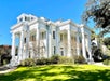 A large white mansion with tall columns and decorative trim stands on a well-manicured lawn under a clear blue sky.