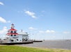 A white building with a red-roofed lighthouse stands by the water under a blue sky with scattered clouds, with grassy foreground and boats docked nearby.