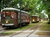 Three green streetcars travel along tracks lined with grass and trees, with passengers visible inside. The front streetcar displays "St. Charles" as its route.