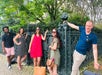 Five people pose casually in front of an ornate iron fence and lush greenery on a sunny day. Some are smiling, and a large tote bag sits on the ground in front of them.