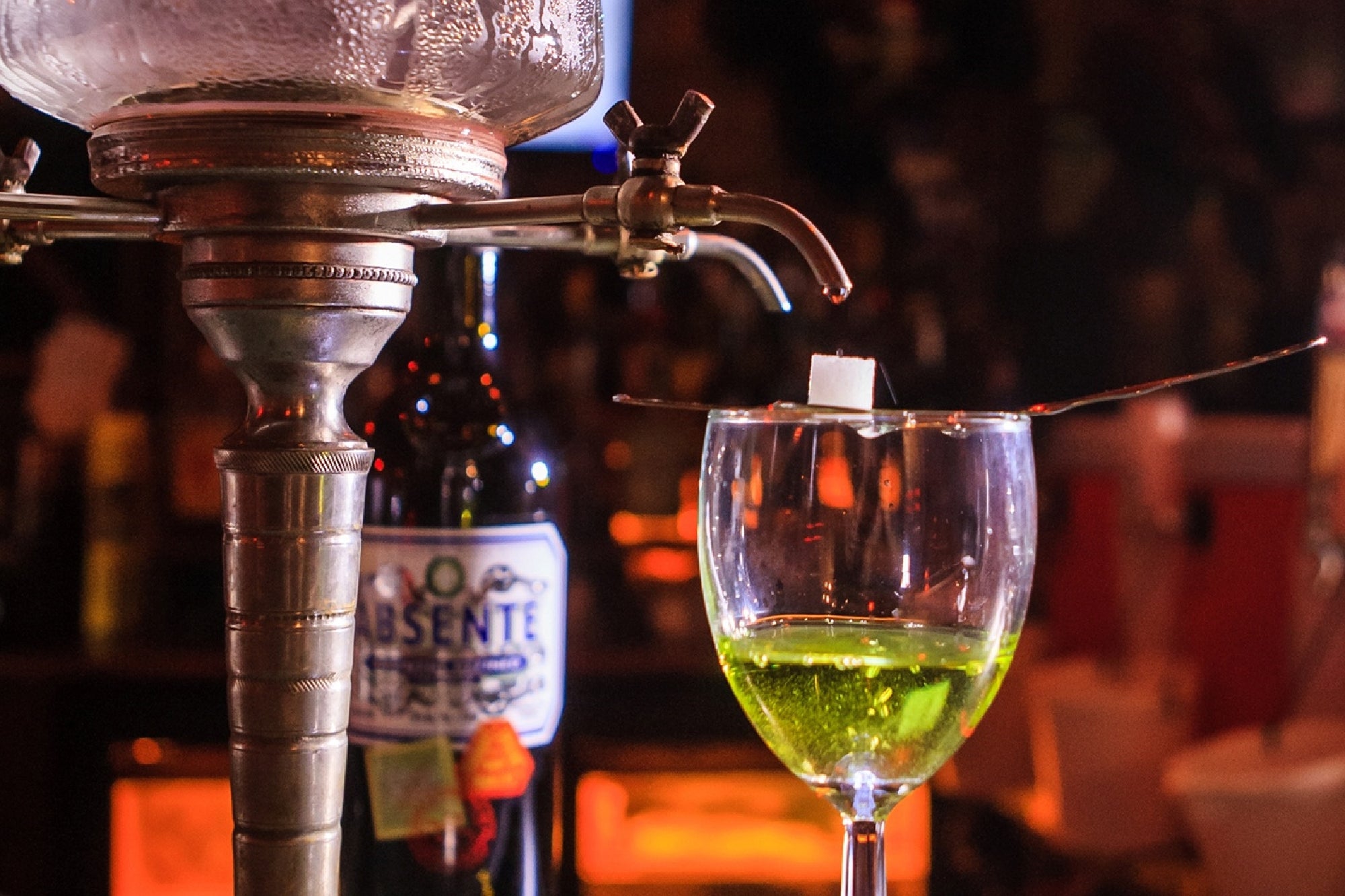 A glass of green absinthe with a sugar cube on a slotted spoon, beside an absinthe fountain and a bottle labeled "Absente.