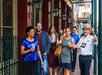 A group of people walk along a city sidewalk, some holding drinks, while a man in a hat gestures and speaks, possibly leading a tour.