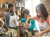 Four people wearing aprons cook together and smile in a kitchen classroom setting with shelves and ingredients in the background.