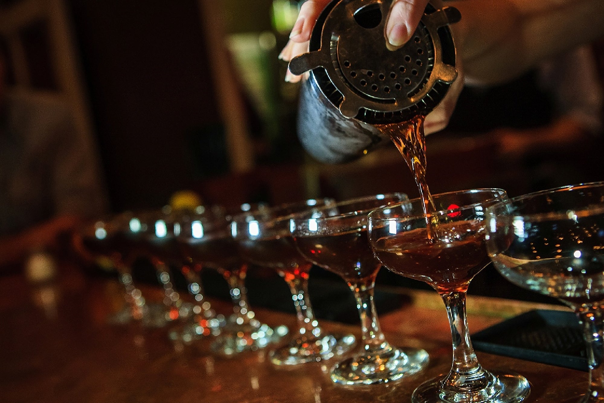 A bartender pours a dark cocktail from a shaker through a strainer into a row of coupe glasses lined up on a bar counter.