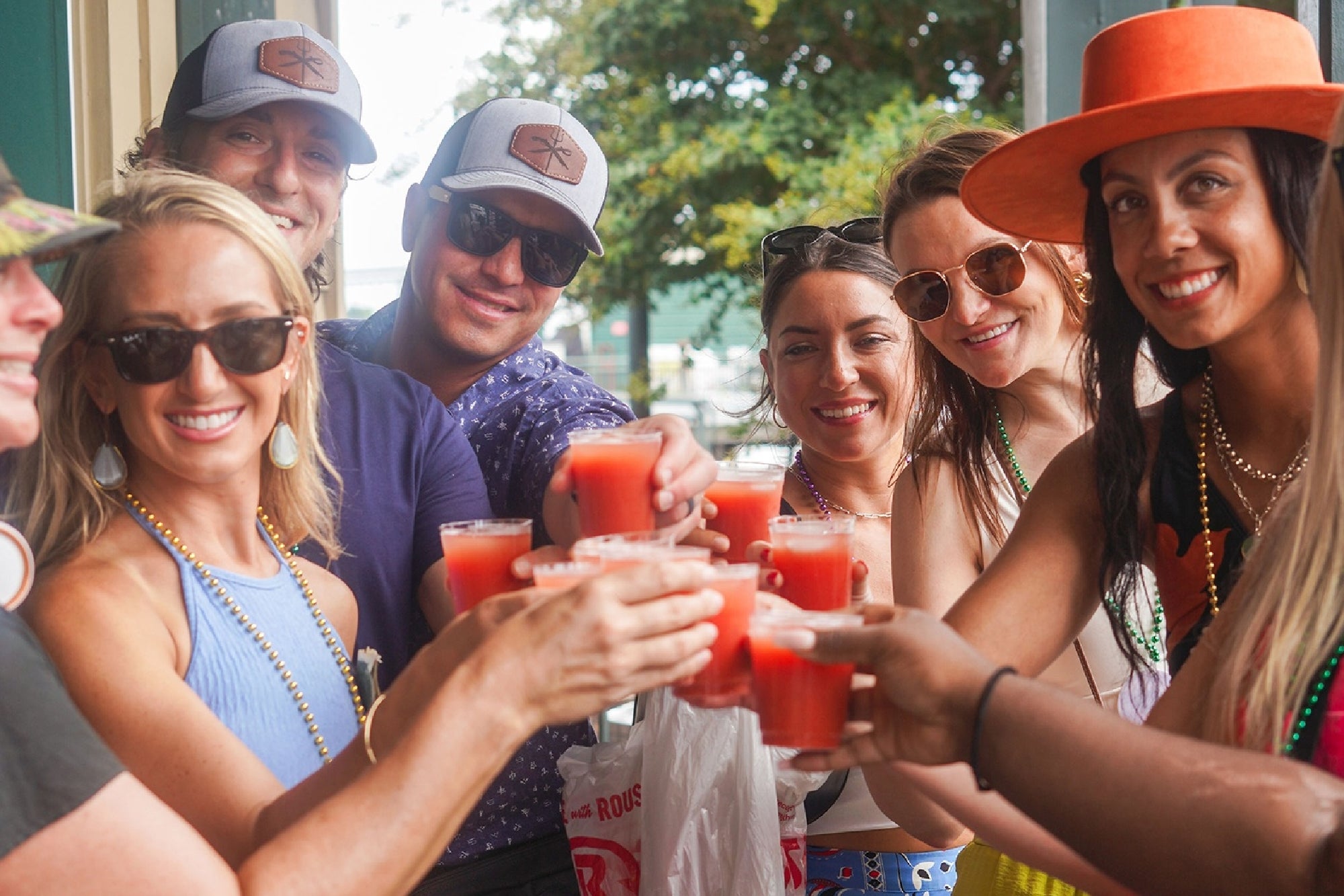 A group of smiling people wearing hats and sunglasses holds up red drinks for a toast while standing outside near greenery.