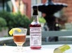 A glass of cocktail garnished with a lemon twist sits next to a bottle of Peychaud's Aromatic Cocktail Bitters on an outdoor table with a fountain in the background.
