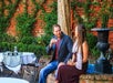 A man and woman sit by a brick wall and fountain in an outdoor patio, holding drinks and talking. Tables with white tablecloths and chairs are nearby.