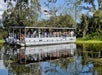 A group of people ride on a covered tour boat over a calm, reflective body of water surrounded by trees and vegetation.