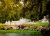 A cemetery with white tombs and grave markers is situated near dense trees and water, with rocks and greenery in the foreground.