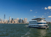 A large white cruise boat in the water with New York City in the back ground on a sunny day with a bright blue sky overhead.