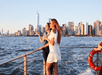 Two young women stranding near at the railing on the top deck of a cruise boat with New York City in the background and the sun shining on them.,