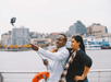 A man holding a selfie stick posing with a woman near the railing of a cruise boat with New York City in the background on a sunny day.