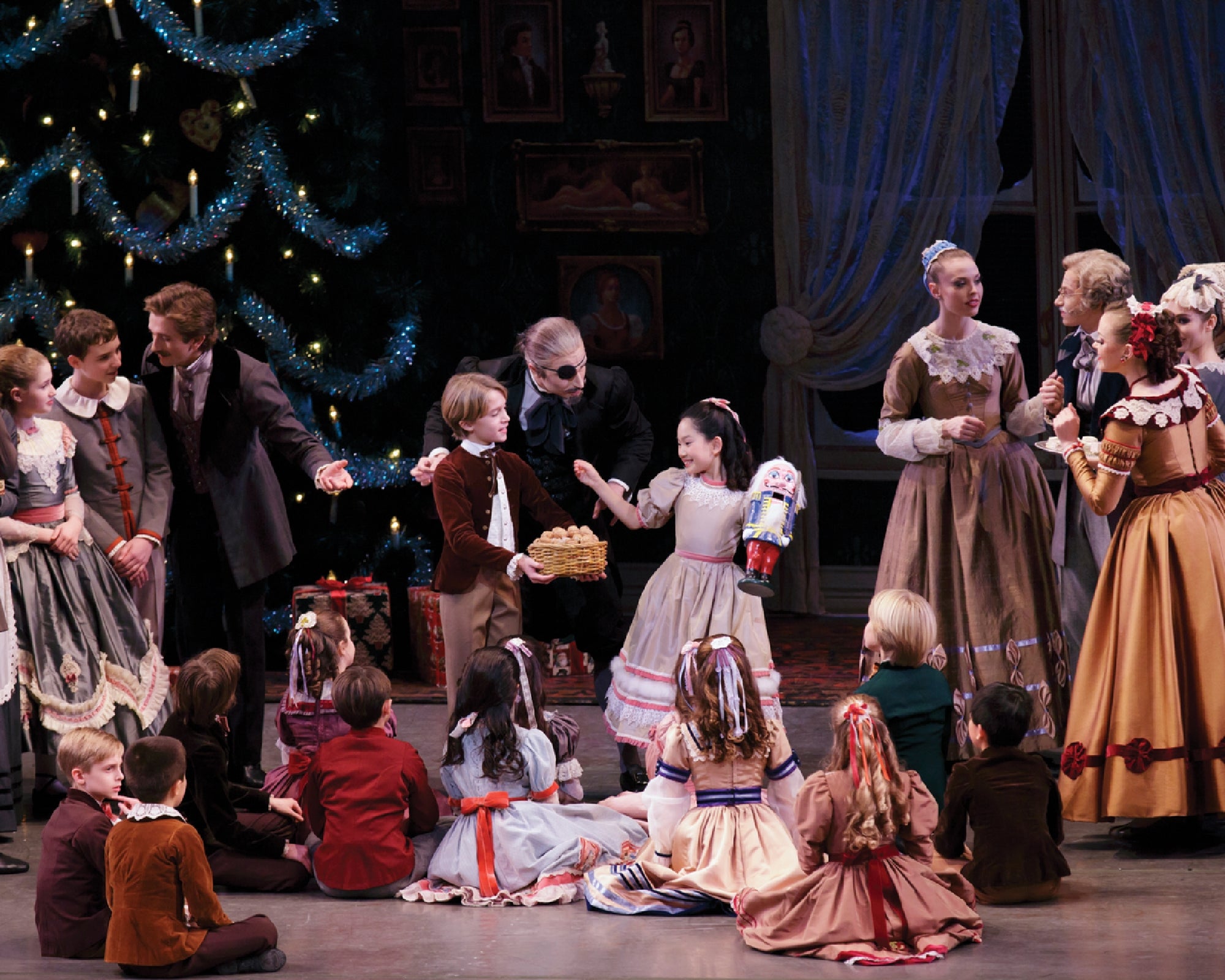 A group of children and adults in period costumes gather around a Christmas tree, exchanging gifts and celebrating in a festive setting.