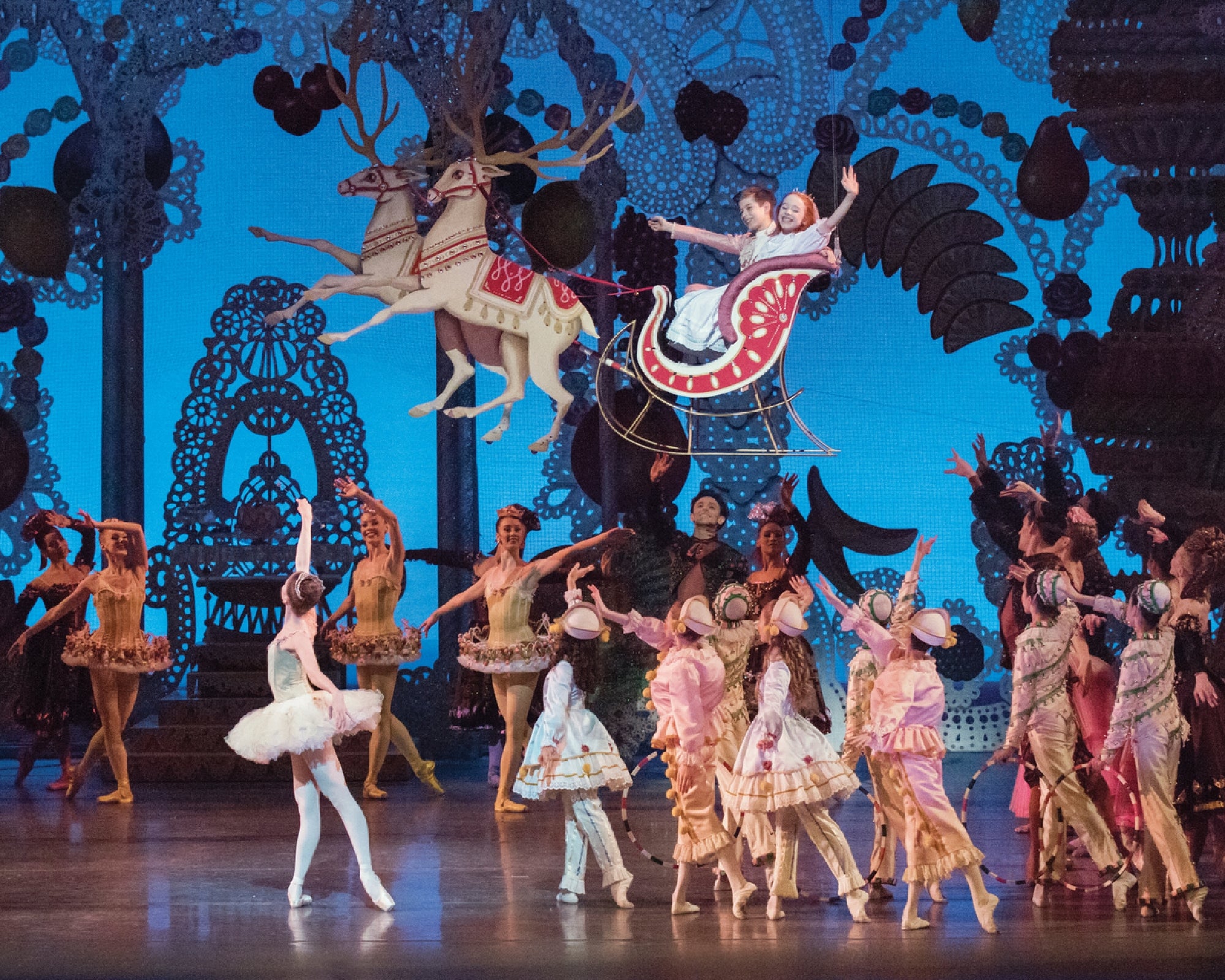 Ballet dancers perform on stage with a backdrop of reindeer pulling a sleigh, surrounded by ornate decorations and a group of performers in colorful costumes.