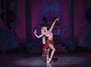 Ballet dancer in ornate costume performs on stage, striking a graceful pose with arms extended, against a decorative backdrop.