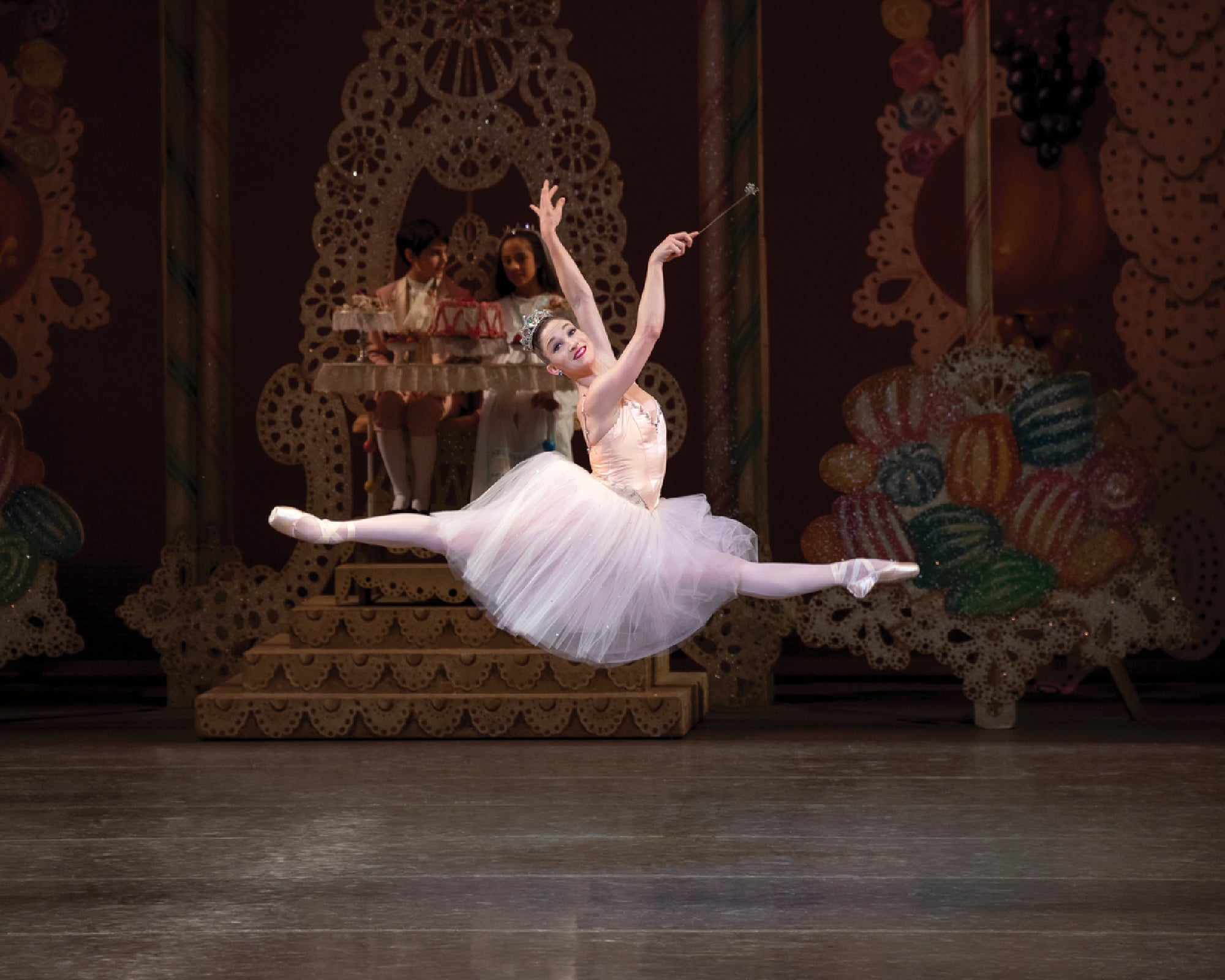 Ballet dancer in mid-leap on stage, wearing a white tutu. The background features elaborate, candy-themed decorations.