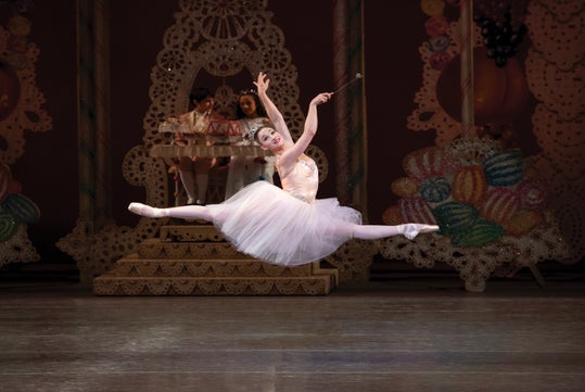 Ballet dancer in mid-leap on stage, wearing a white tutu. The background features elaborate, candy-themed decorations.