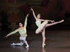 Two ballet dancers perform on stage. The male dancer kneels while the female dancer balances on one leg, extending the other gracefully. They wear elaborate costumes.