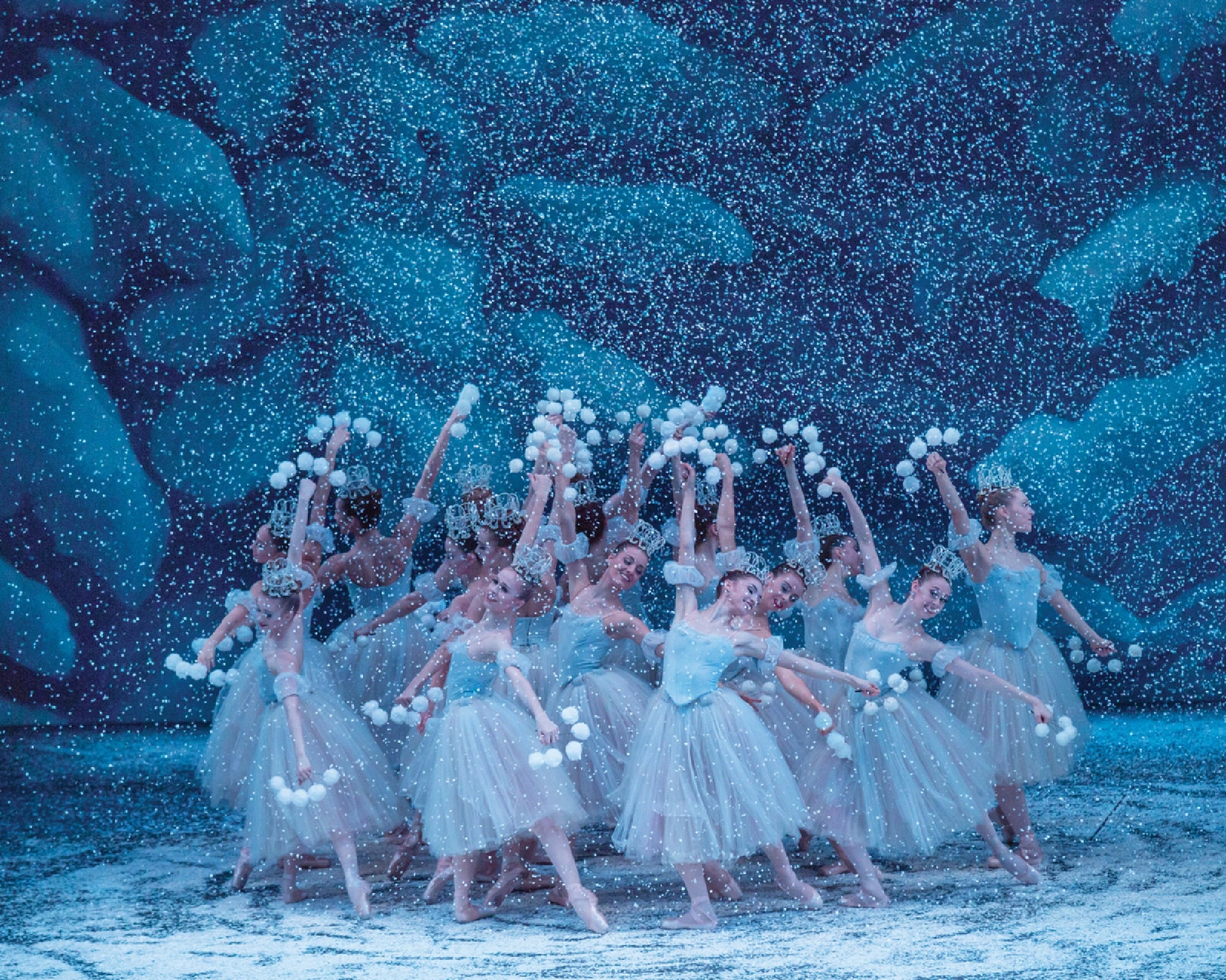 Ballet dancers in blue dresses perform on a snowy stage, holding pom-pom props against a backdrop of snow-covered trees.