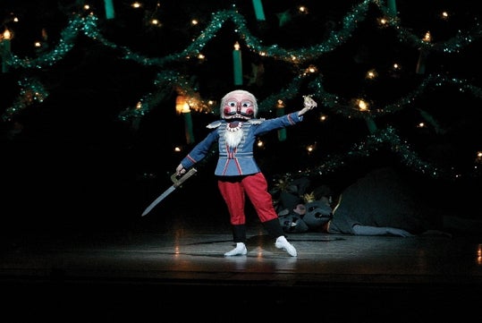 A performer in a nutcracker costume stands on stage holding a sword, with a large mouse figure lying on the floor in the background, and a decorated tree with lights behind them.