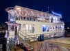 A large, brightly lit two-story boat is docked at night, with people boarding via a ramp from a wooden pier lined with red rope barriers.