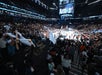 A large crowd in an indoor basketball arena cheers and waves towels while a game is played on the court; a scoreboard and jumbo screen hang above the action.
