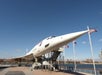 A retired supersonic Concorde jet is displayed outdoors on a platform by the waterfront, with American flags visible and a clear blue sky overhead.