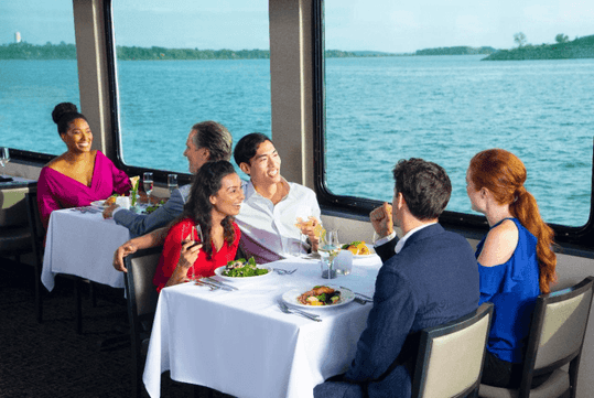 A group of people at two tables on a boat enjoy food and drinks. Large windows offer a view of the water and distant land.